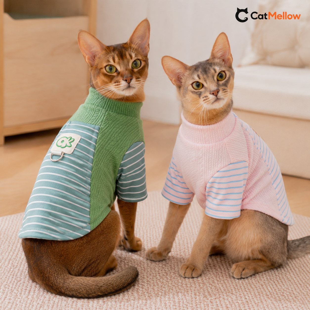 Two cats wearing the green and pink CatMellow pastel stripe knit shirts in a warm home.