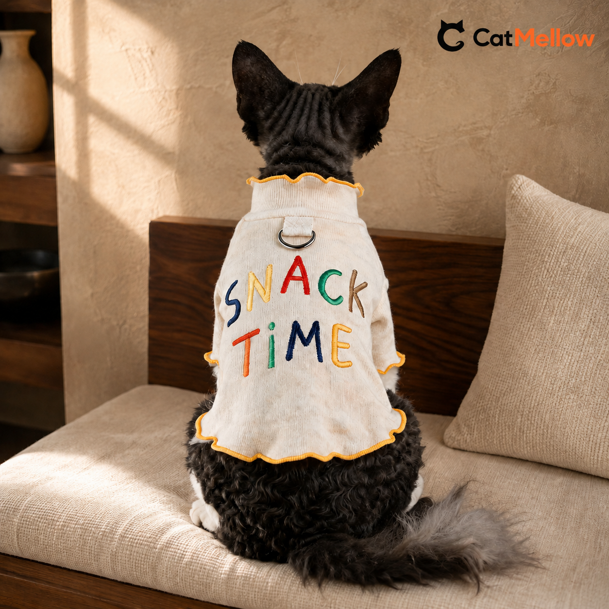 Back view of a black-and-white cat wearing the cream CatMellow Snack Time embroidered T-shirt on a walnut bench